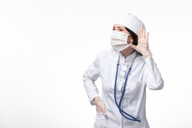Dottoressa vista frontale in tuta medica bianca sterile con maschera a causa del coronavirus che cerca di ascoltare sulla malattia del muro bianco covid - malattia da virus pandemico
