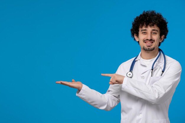 Dottori giorno ricci bel ragazzo carino in uniforme medica che punta a sinistra e sorridente