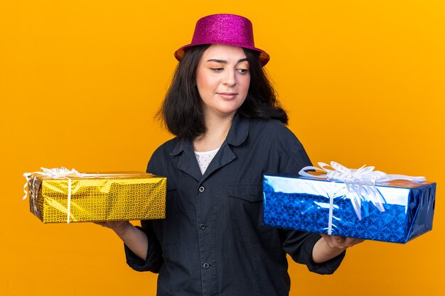 Dubbiosa giovane ragazza caucasica che indossa un cappello da festa con pacchetti regalo guardando uno di loro isolato sulla parete arancione