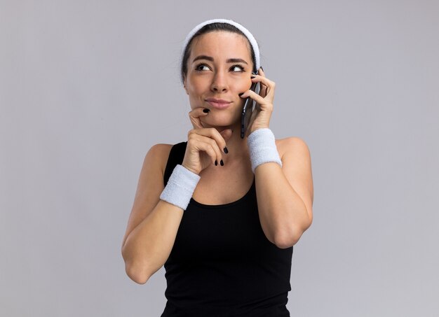 Dubbiosa giovane ragazza piuttosto sportiva che indossa fascia e braccialetti che parla al telefono tenendo la mano sul mento guardando in alto