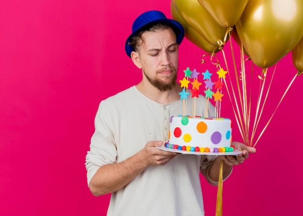 Dubbioso giovane bel partito slavo ragazzo indossa il cappello del partito che tiene palloncini e torta di compleanno con le stelle guardando la torta isolata sulla parete rosa con lo spazio della copia