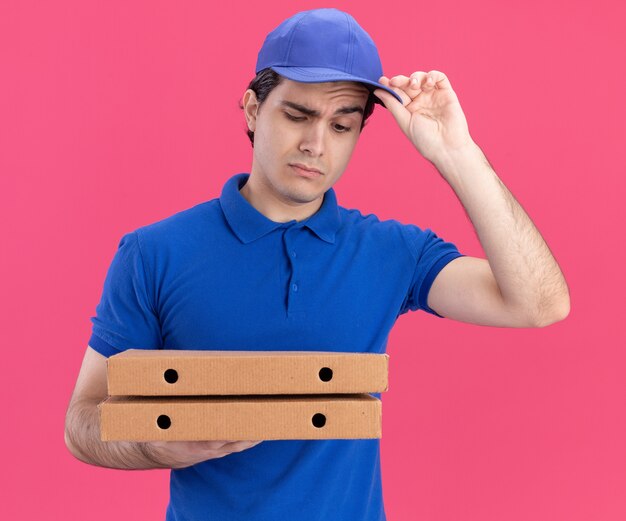 Dubbioso giovane fattorino caucasico in uniforme blu e berretto che tiene e guarda i pacchetti di pizza afferrando il berretto isolato sul muro rosa