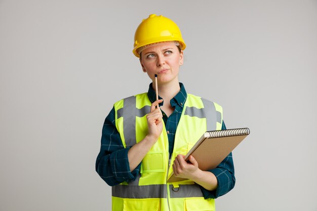 Dubbioso giovane operaio edile femminile che indossa il casco di sicurezza e giubbotto di sicurezza che tiene il blocco note che tocca il mento con la matita che osserva in su