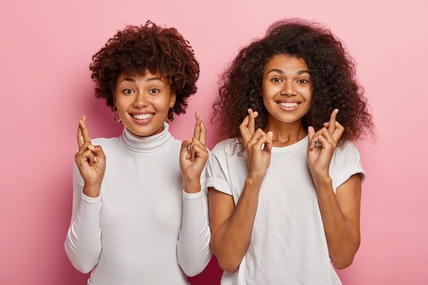 Due allegre donne dai capelli ricci tengono le dita incrociate per buona fortuna, vestite in abito bianco, sorridono felicemente, anticipano i risultati degli esami, stanno su sfondo rosa.