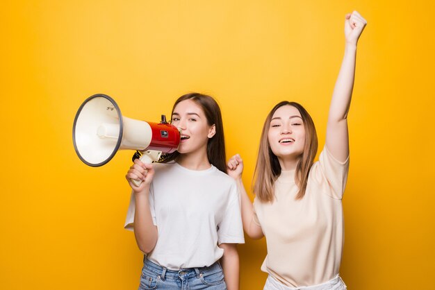 Due amiche irritate delle ragazze delle giovani donne urlano nel megafono isolato sulla parete gialla. Persone concetto di stile di vita. Mock up copia spazio.