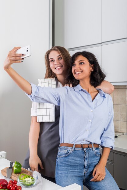 Due amici femminili che prendono selfie sul telefono cellulare in cucina