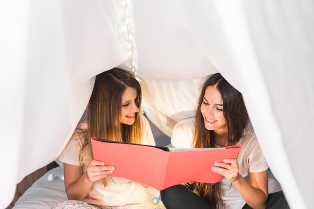 Due amici femminili felici che esaminano l&#39;album di foto che si siede sul letto sotto la tenda bianca