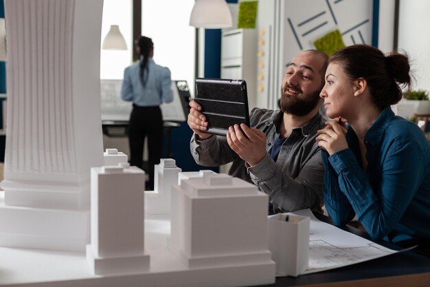 Due architetti che lavorano insieme guardando tablet con progetti seduti alla scrivania in un moderno ufficio architettonico. Ingegneri di progetto che lavorano in team utilizzando un dispositivo digitale accanto al tavolo con un modello in scala.