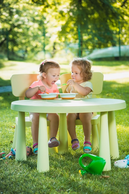 Due bambine che si siedono ad una tavola e che mangiano insieme contro il prato inglese verde