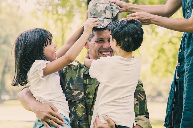 Due bambini felici e la loro mamma incontrano e abbracciano il papà militare in uniforme mimetica all'aperto
