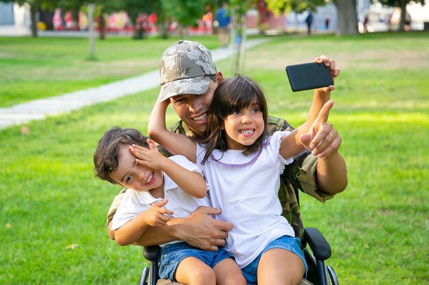 Due bambini felici seduti sulle ginocchia di papà e prendendo selfie sul cellulare. Uomo militare disabile che cammina con i bambini nel parco. Veterano di guerra o concetto di disabilità