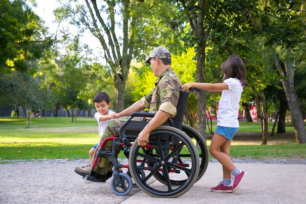 Due bambini piccoli che camminano con papà disabili militari in sedia a rotelle nel parco cittadino. Vista laterale. Veterano di guerra o concetto di disabilità
