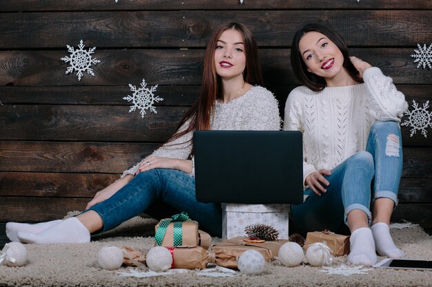 Due belle donne giacciono sul pavimento con un laptop, tra i regali di Natale