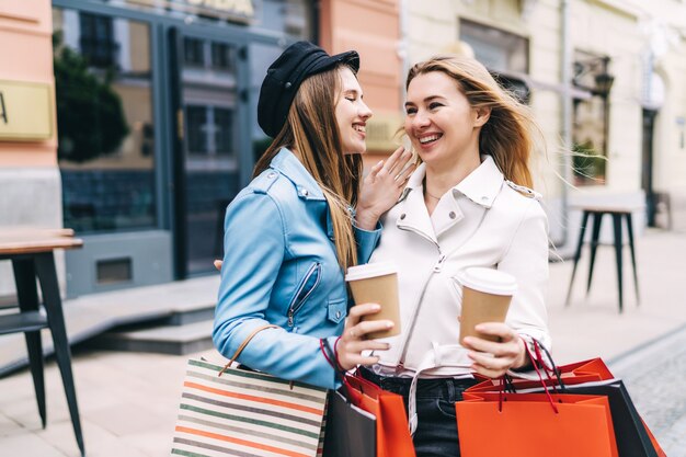 Due belle donne in mezzo alla strada con caffè e borse della spesa in mano sono in piedi e discutono con interesse