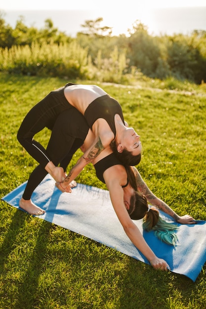 Due belle donne in top sportivi neri e leggings che allenano lo yoga posano insieme sul tappetino Giovani donne che praticano yoga sull'erba all'aperto