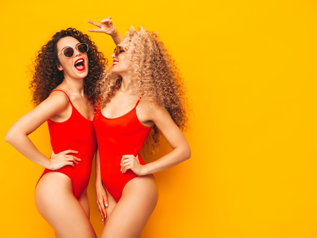 Due belle donne sexy hipster sorridenti in costumi da bagno costumi da bagno estivi rossi Modelli alla moda con acconciatura di riccioli afro divertendosi in studio Femmina calda isolata su giallo in occhiali da sole