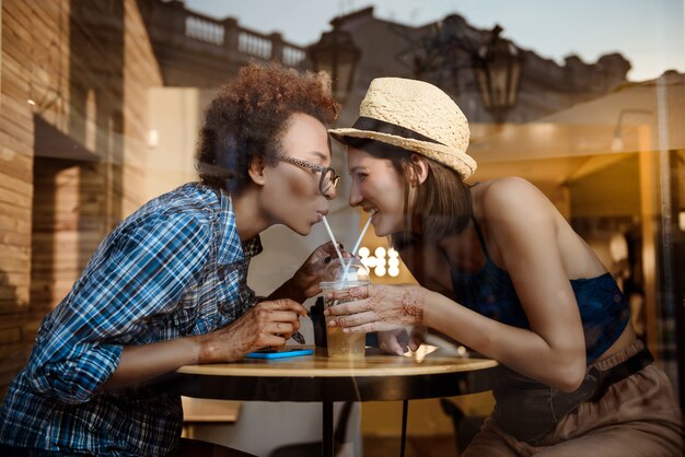 Due belle ragazze che sorridono, bevendo dai tubi, riposando nella caffetteria. Tiro esterno.