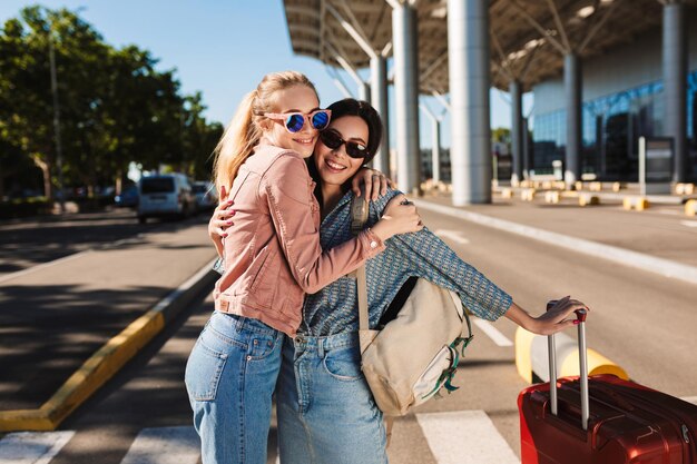 Due belle ragazze in occhiali da sole che si abbracciano felicemente mentre guardano a porte chiuse con la valigia rossa e lo zaino sulla spalla all'aperto vicino all'aeroporto
