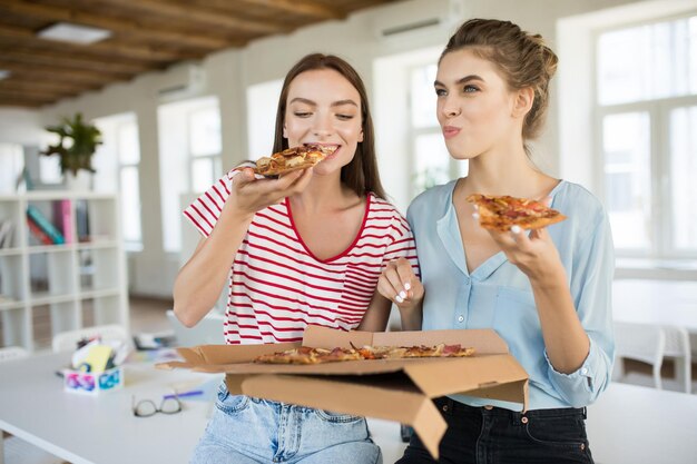 Due belle ragazze sedute sulla scrivania mangiano con gioia una grande pizza mentre trascorrono del tempo insieme in un ufficio moderno