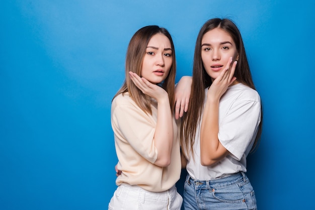 Due belle sorprese che urlano con le mani alzate, vestite in abiti casual su una parete blu. Concetto di emozioni di persone.