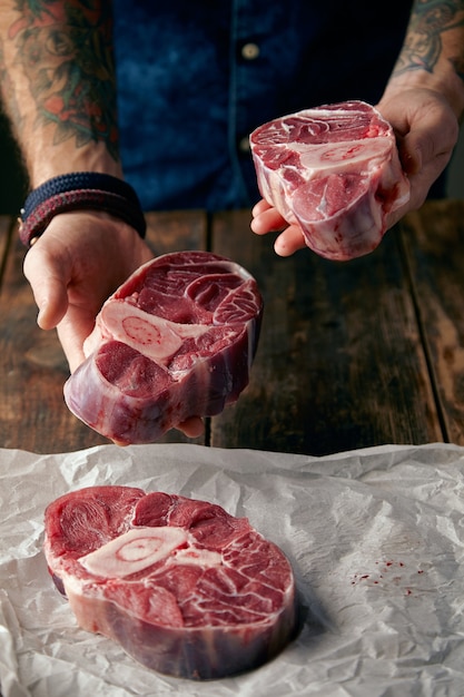 Due bistecche in mani tatuate e altre su carta artigianale. Offriti di comprare carne sulla macchina fotografica.