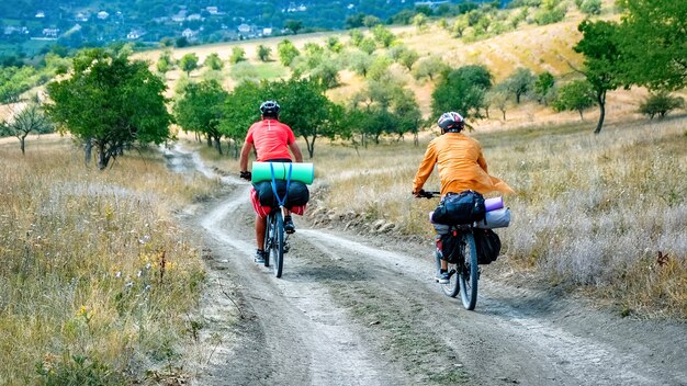 Due ciclisti in caschi con biciclette piene di roba da viaggiatore che si muovono sulla strada di campagna tra rari alberi verdi