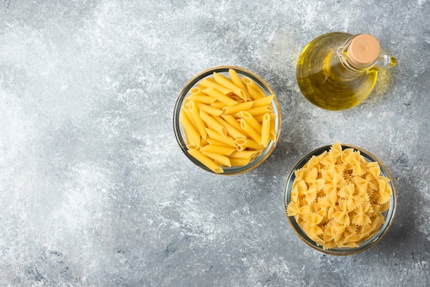 Due ciotole di pasta cruda di penne e farfalle con una bottiglia di olio d'oliva su fondo di marmo.