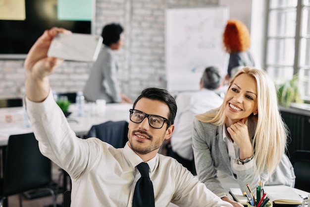 Due colleghi che si divertono in ufficio e si fanno selfie con il cellulare Ci sono persone sullo sfondo