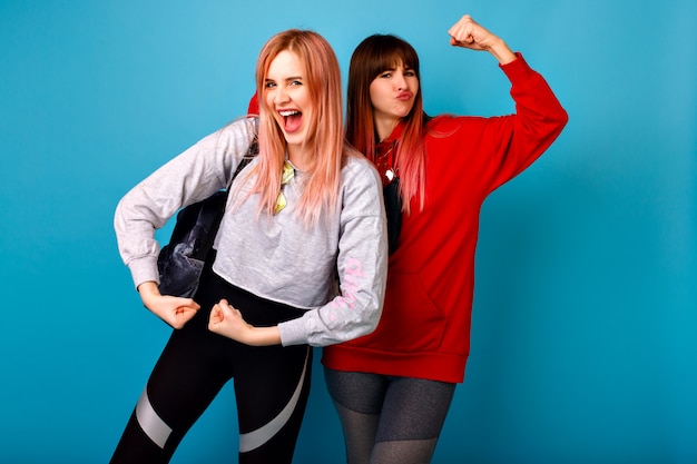 Due divertenti hipster giovane donna che mostra bicipiti, parete blu, vestiti sportivi fitness, emozioni uscite, coppia che impazzisce insieme.