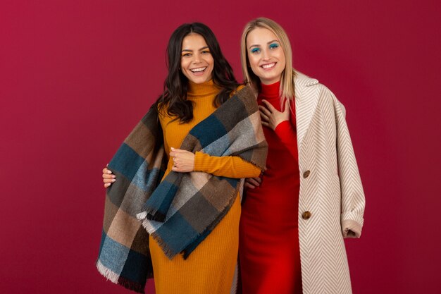 Due donne alla moda attraenti sorridenti in vestito da modo di autunno inverno e posa del cappotto isolata sulla parete rossa