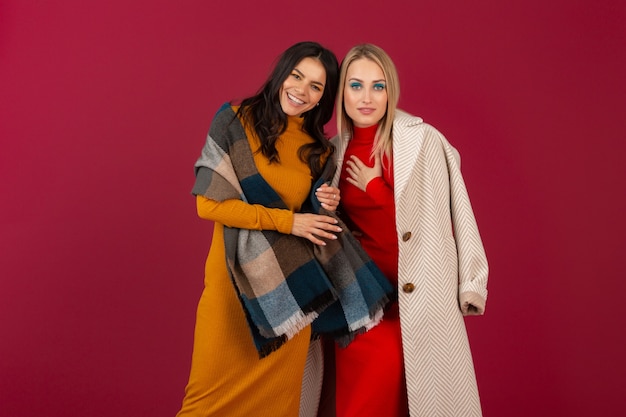 Due donne alla moda attraenti sorridenti in vestito da modo di autunno inverno e posa del cappotto isolata sulla parete rossa