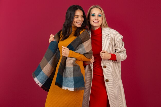 Due donne alla moda attraenti sorridenti in vestito da modo di autunno inverno e posa del cappotto isolata sulla parete rossa