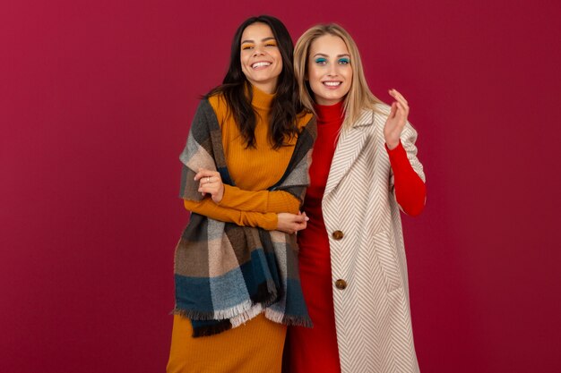 Due donne alla moda attraenti sorridenti in vestito da modo di autunno inverno e posa del cappotto isolata sulla parete rossa