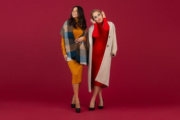 Due donne alla moda in abito da moda autunno inverno e cappotto in posa isolato sulla parete rossa