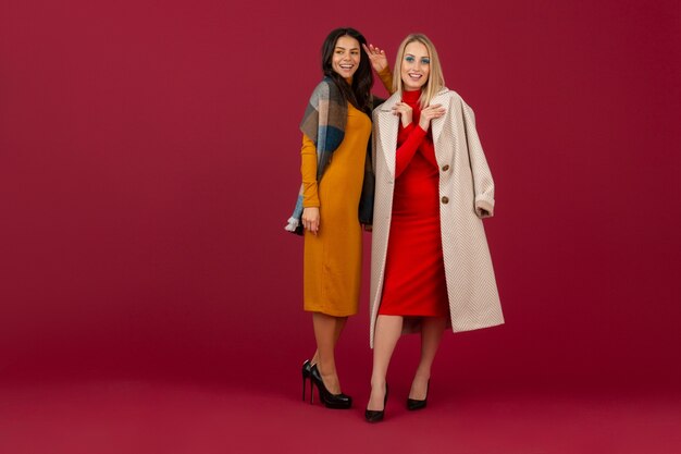 Due donne alla moda in abito da moda autunno inverno e cappotto in posa isolato sulla parete rossa