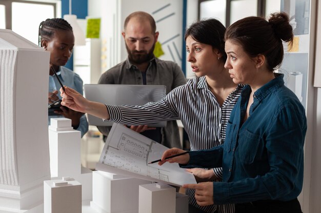 Due donne architetti professionisti in possesso di progetti accanto a colleghi in possesso di laptop guardando i piani di costruzione. Gruppo di ingegneri architettonici focalizzati che analizzano il modello in scala di schiuma bianca.