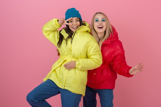 Due donne attive attraenti in posa sul muro rosa in piumino invernale colorato di colore rosso e giallo brillante, amici che si divertono insieme, tendenza moda cappotto caldo, facce buffe pazze