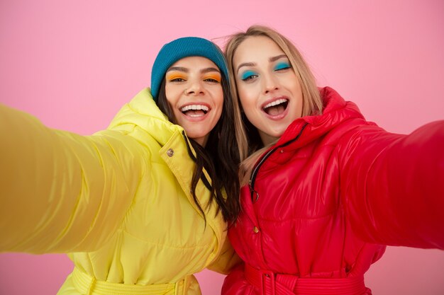 Due donne attraenti in posa su sfondo rosa in piumino invernale colorato di colore rosso e giallo brillante, amici che si divertono insieme, tendenza di moda vestiti caldi, prendendo selfie