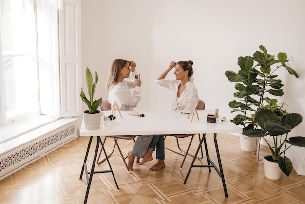 Due donne caucasiche bionde che eseguono la procedura di lifting mentre si siedono a tavola in una stanza luminosa. Concetto di cura della pelle e giornata della salute