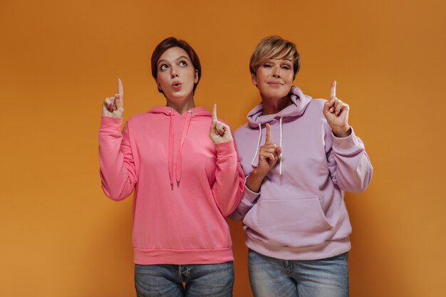 Due donne fantastiche con i capelli corti in larghe felpe con cappuccio rosa e lilla e jeans che mostrano i pollici in su e posano su sfondo arancione.