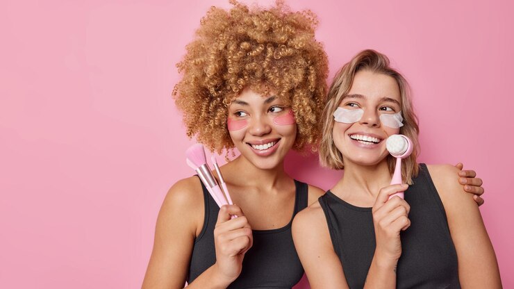 Foto gratuito due donne felici applicano i cerotti di bellezza usano i pennelli cosmetici e la spazzola per il viso per prendersi cura di se stessa sottoporsi a routine quotidiane di coccole abbracciare guardare con gioia posare su sfondo rosa