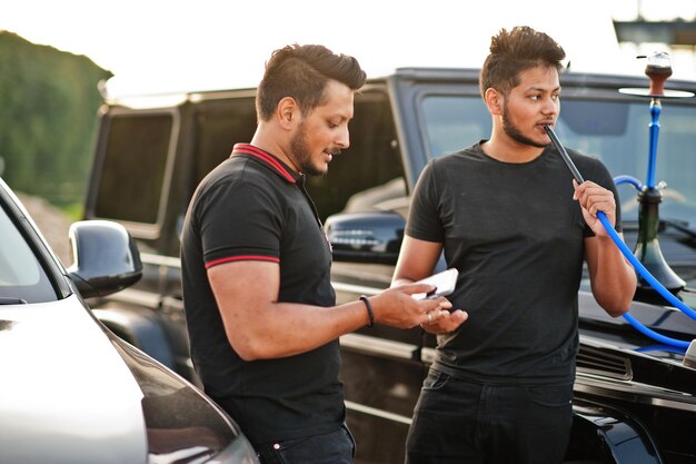 Due fratelli asiatici indossano abiti neri in posa vicino a suv e fumano narghilè