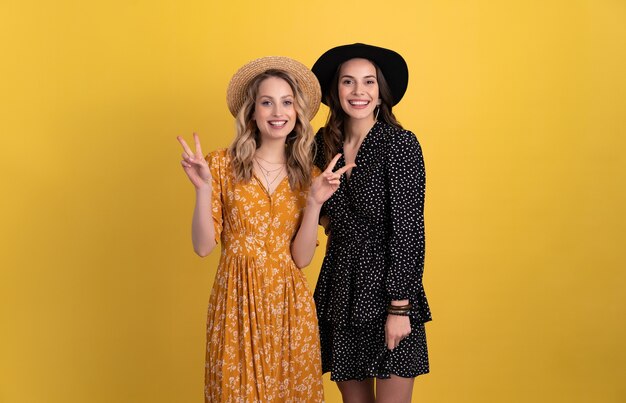 due giovani belle donne amiche insieme isolate su giallo in abito nero e giallo e cappello elegante boho divertendosi