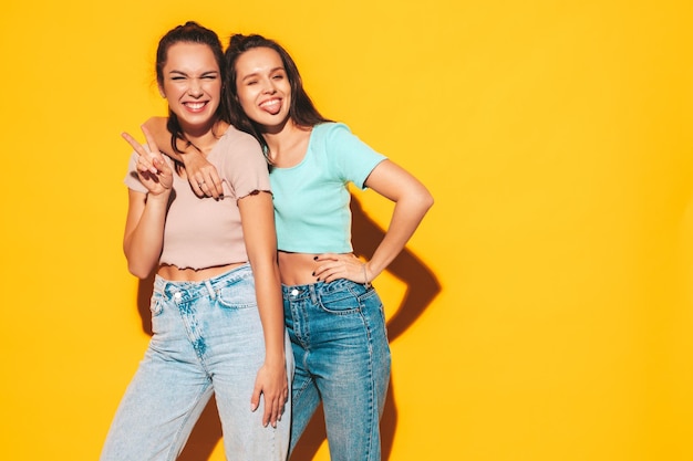 Due giovani belle donne bionde sorridenti hipster in abiti estivi alla moda Donne spensierate sexy che posano vicino al muro giallo in studio Modelli positivi che si divertono Allegri e felici