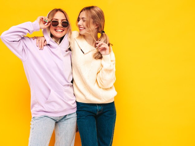 Due giovani belle donne bionde sorridenti hipster in abiti estivi alla moda Donne spensierate sexy che posano vicino al muro giallo in studio Modelli positivi che si divertono Allegri e felici