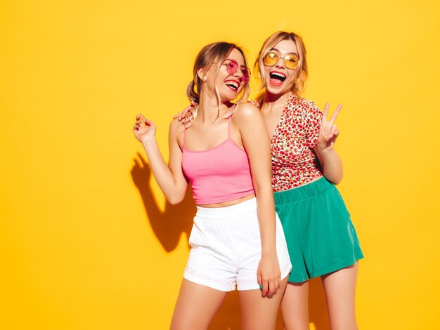 Due giovani belle donne bionde sorridenti hipster in abiti estivi alla moda Donne spensierate sexy in posa vicino al muro giallo in studio Modelli positivi che si divertono Allegri e felici In occhiali da sole