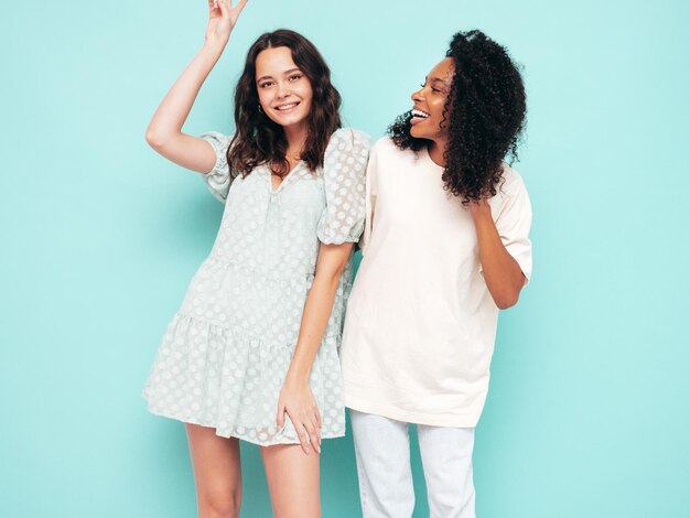 Due giovani belle donne hipster internazionali sorridenti in abiti estivi alla moda Donne spensierate sexy in posa vicino al muro blu in studio Modelli positivi divertendosi Concetto di amicizia