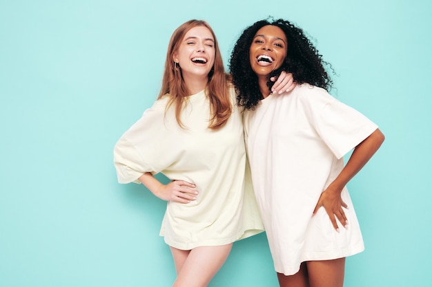 Due giovani belle donne hipster internazionali sorridenti in abiti estivi alla moda Donne spensierate sexy in posa vicino al muro blu in studio Modelli positivi divertendosi Concetto di amicizia