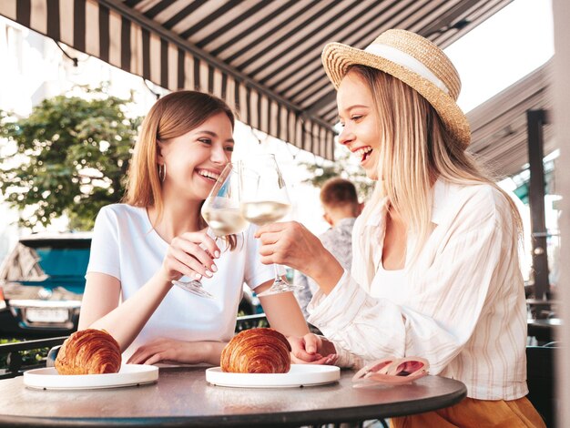 Due giovani belle donne hipster sorridenti in abiti estivi alla moda Donne spensierate che posano al caffè sulla veranda in stradaModelli positivi che bevono vino bianco Godersi la vacanzaMangiare croissant
