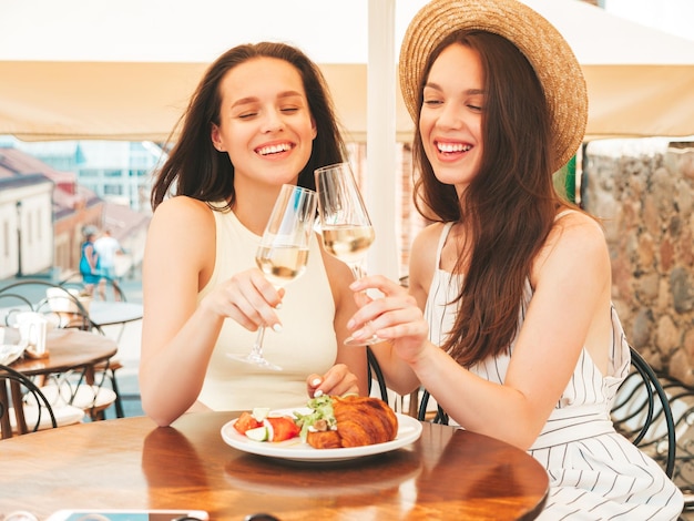 Due giovani belle donne hipster sorridenti in abiti estivi alla moda Donne spensierate che posano al caffè veranda in stradaModelli positivi che bevono vino bianco con cappelloGodersi le vacanze